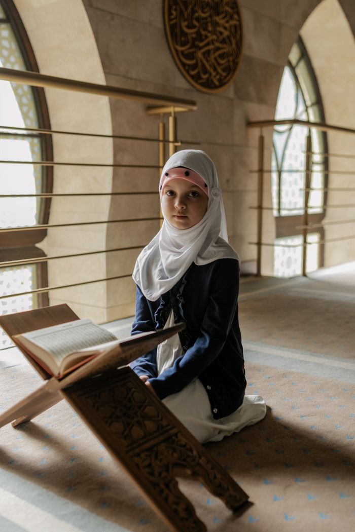 Crafting Captivating Headlines: Your awesome post title goes here A young girl in traditional attire prays with the Quran inside a beautifully lit mosque, reflecting Muslim culture.