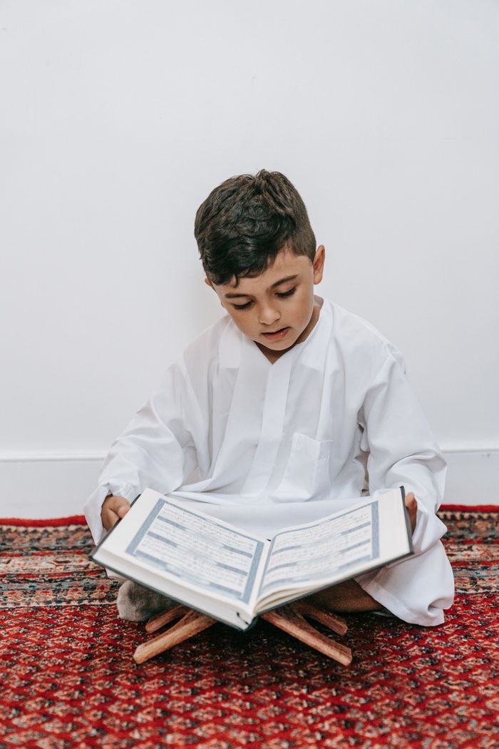 A child sits cross-legged reading a holy book indoors, emphasizing cultural and religious learning.